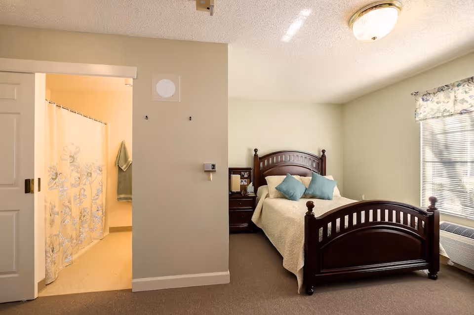 A cozy bedroom in a senior living facility with a wooden bed frame, beige bedding, and two blue pillows. There is a nightstand with a lamp and picture frames next to the bed. A window with floral curtains allows natural light into the room. To the left, an open doorway reveals a bathroom with a shower curtain featuring a floral design and a green towel hanging on the wall.