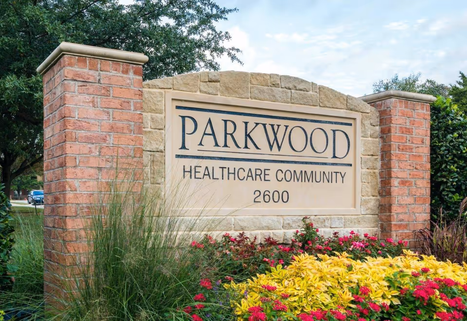 Stone and brick sign for Parkwood Healthcare Community with the number 2600, surrounded by green plants and colorful flowers under a partly cloudy sky.
