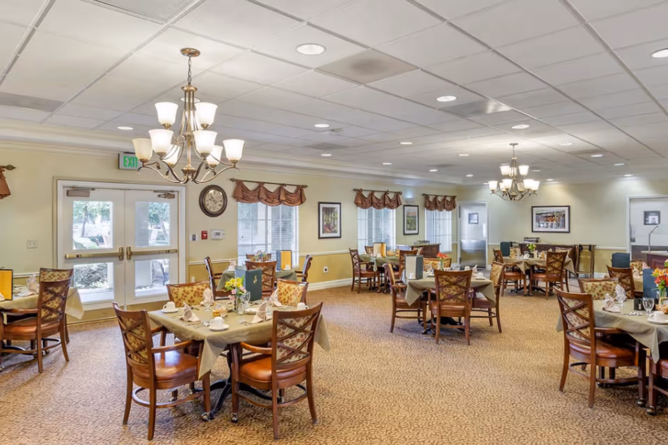 Dining room with several set tables, chandeliers, and chairs in a senior living facility.