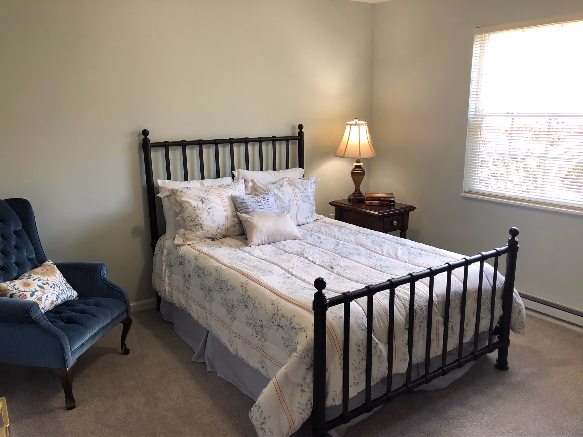 A bedroom with a black metal bed frame, a bed made with floral patterned bedding and multiple pillows. To the right of the bed is a wooden nightstand with a lamp and two books on it. To the left of the bed is a blue upholstered armchair with a floral cushion. A window with blinds is on the right wall, letting in natural light.