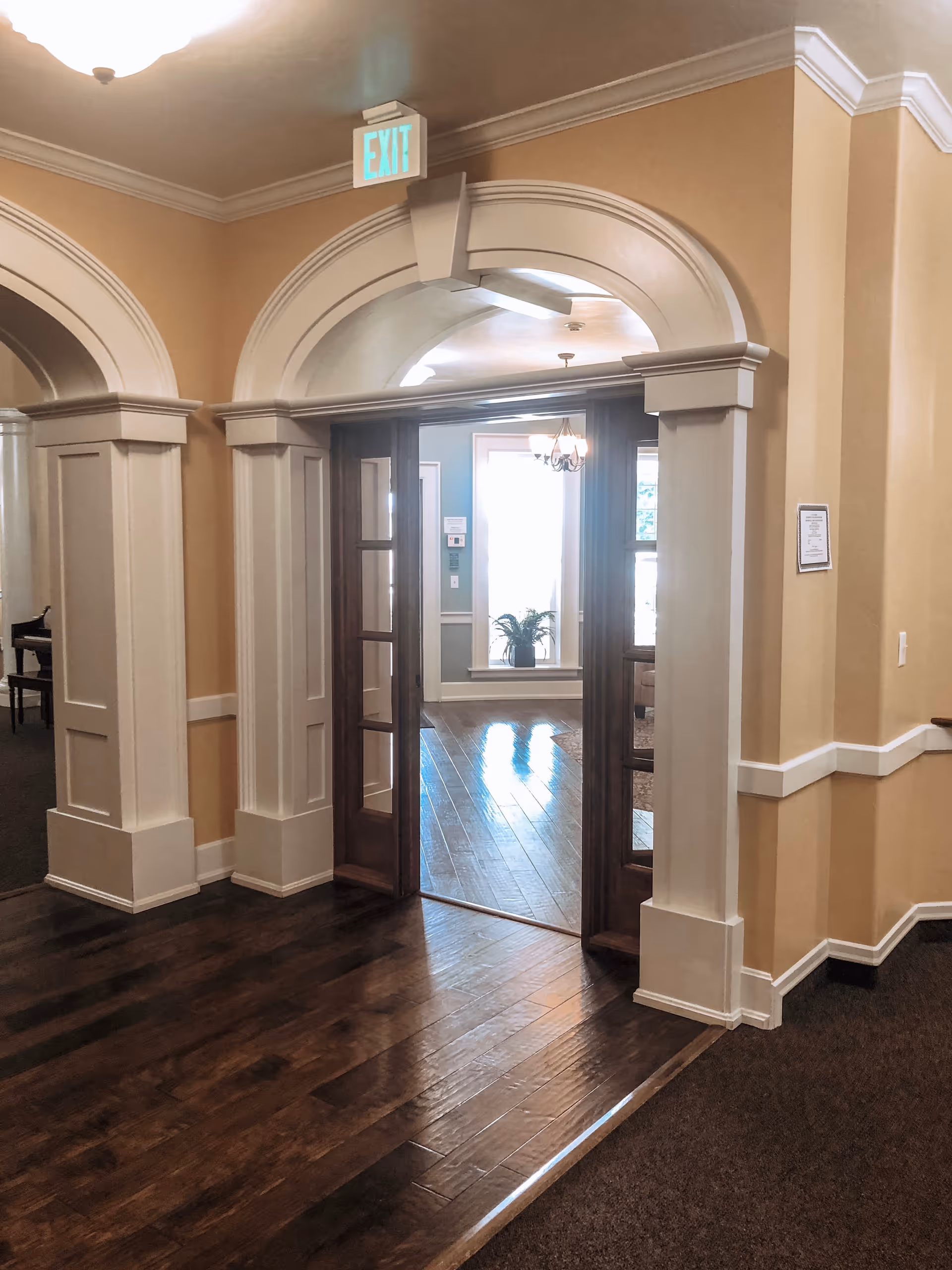 Interior view of a hallway in an assisted living facility with beige walls, white trim, and dark wooden floors. There is an arched doorway with wooden double doors leading to a well-lit room with a chandelier and a potted plant by the window. An exit sign is visible above the doorway.