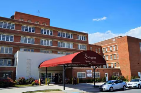 Brick senior living building with a red canopy entrance and parked cars.