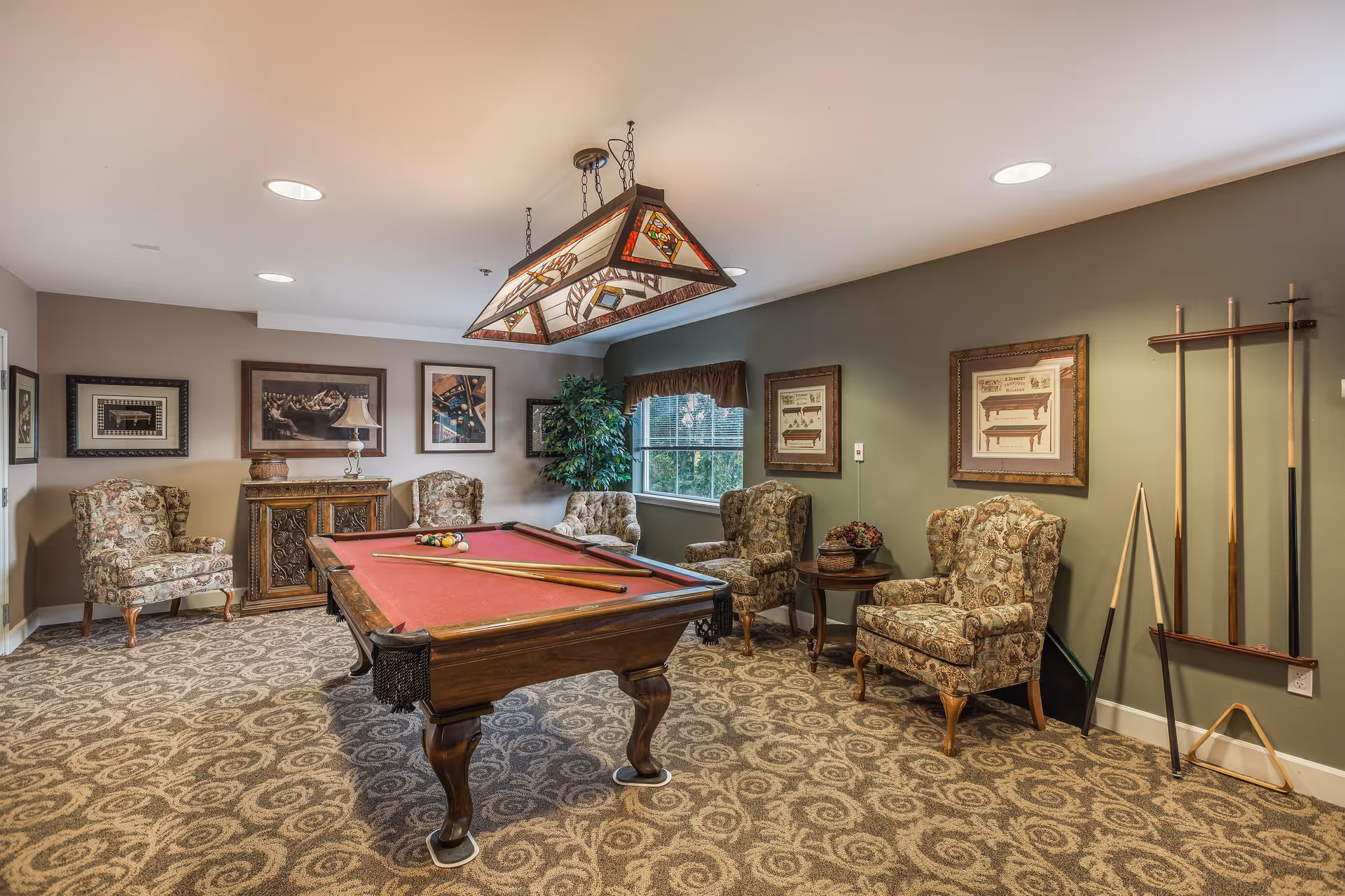 A cozy game room featuring a red felt pool table with cues and balls set up. The room has patterned carpet, four upholstered armchairs with floral designs, framed artwork on the walls, a window with a valance, and a hanging stained glass light fixture above the pool table. A rack holding pool cues is mounted on the wall.