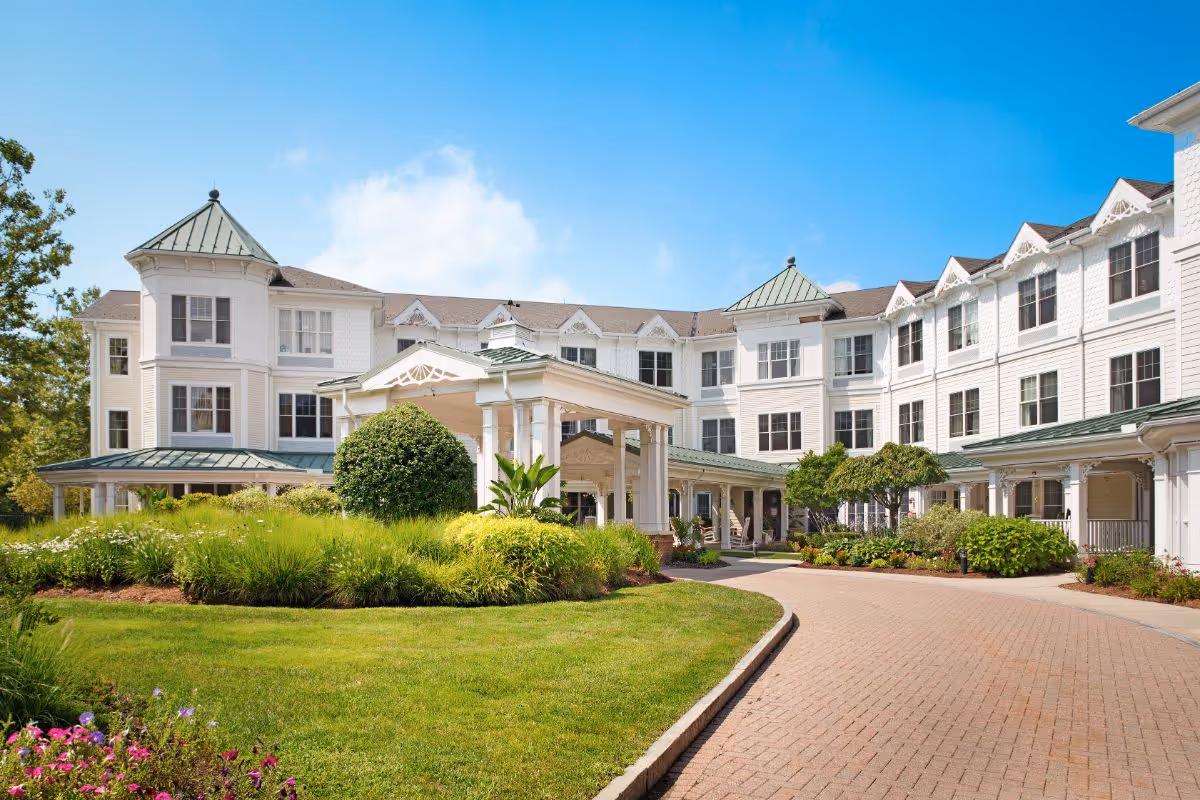 Exterior view of a large, white senior living facility building with multiple windows and green roofs under a clear blue sky. The entrance features a covered driveway with white pillars, surrounded by well-maintained green lawns, bushes, and colorful flowers.