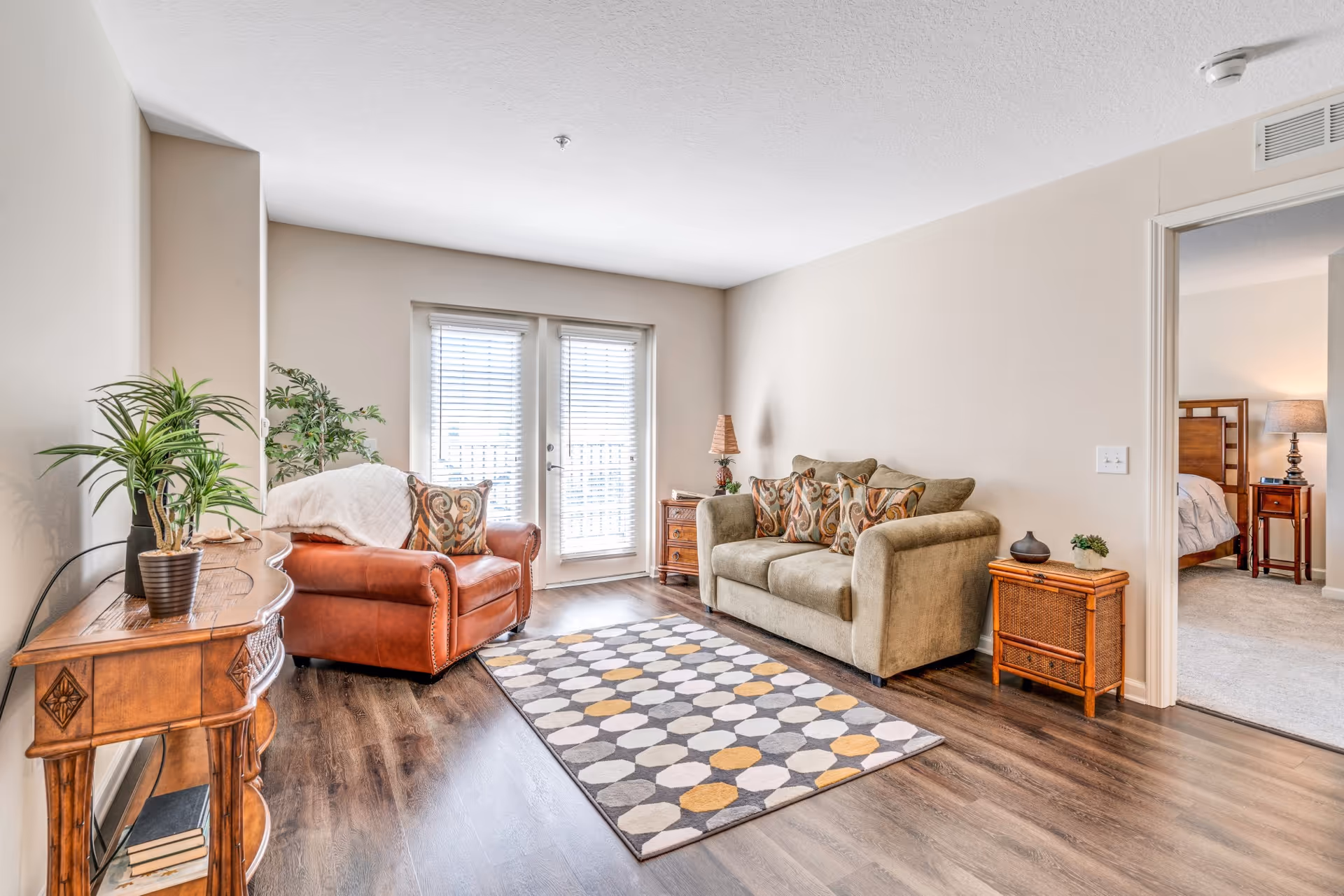 Bright living room with a sofa, leather armchair, patterned rug, wooden side tables, and glass doors letting in natural light.