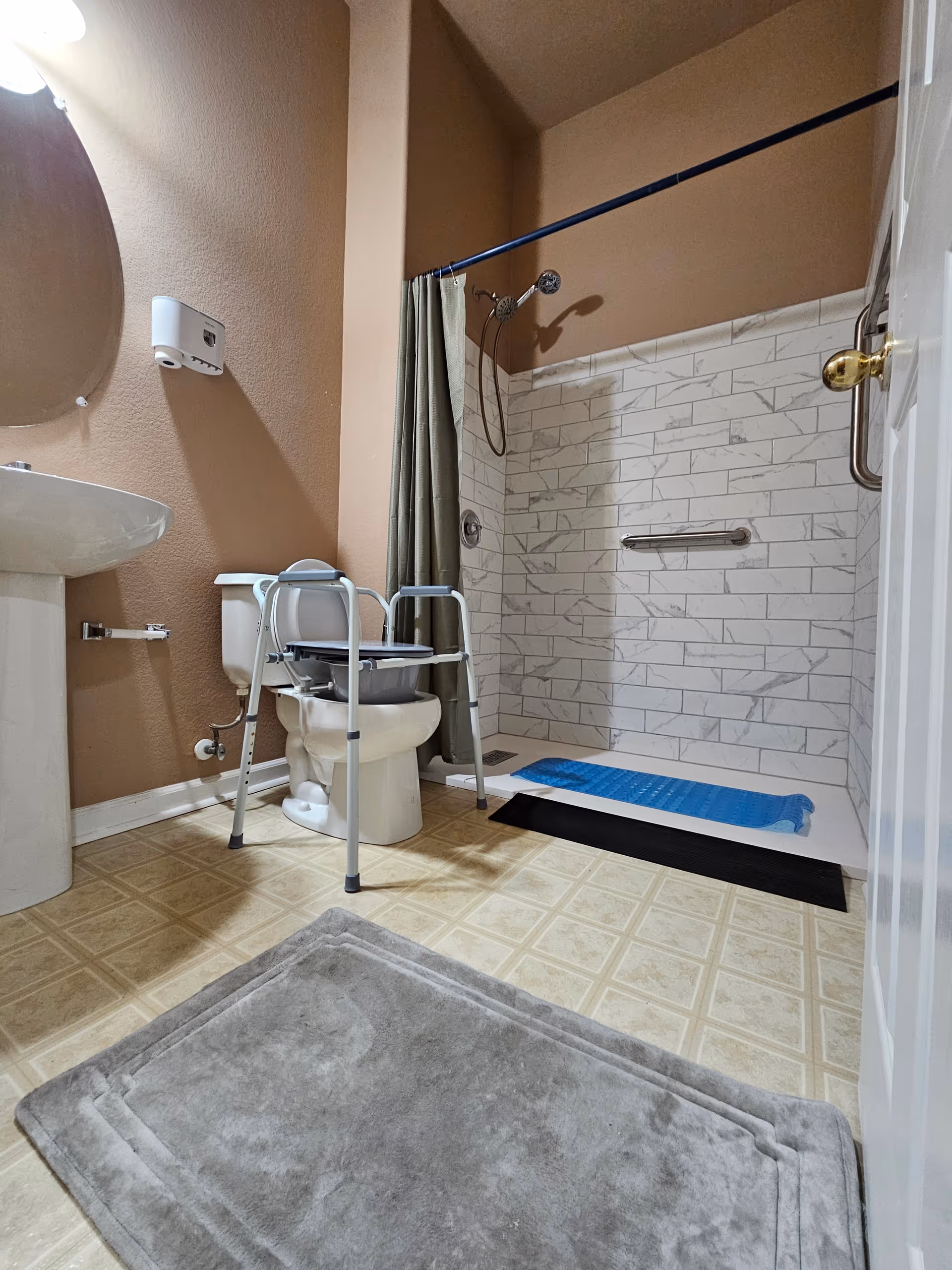 Accessible bathroom showing a toilet with a commode frame, pedestal sink, walk-in tiled shower with grab bar and showerhead, and floor mats.