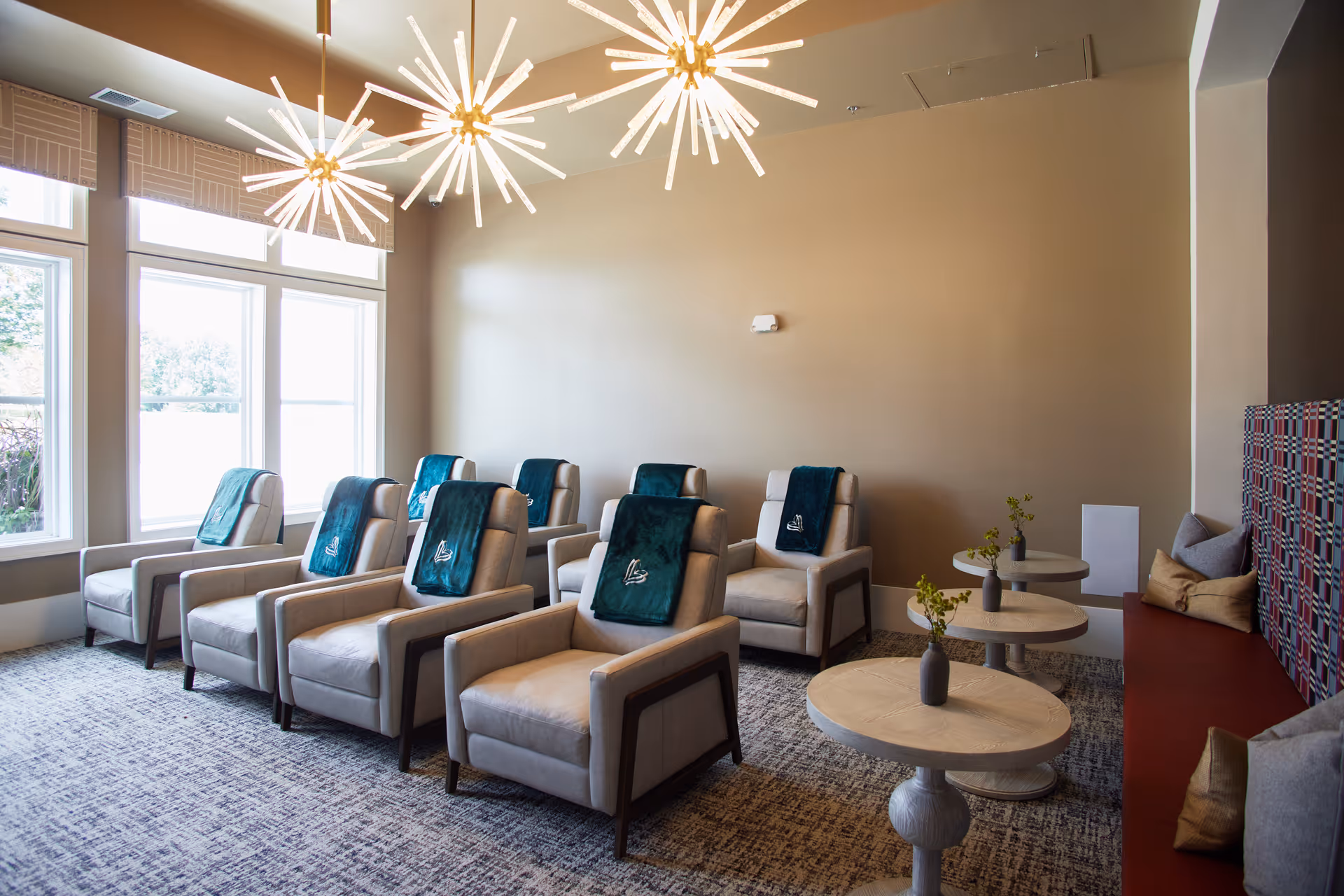 Bright communal lounge with rows of beige recliners each draped with teal throws, round side tables, and modern starburst chandeliers.