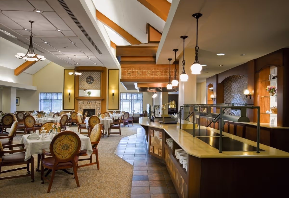 A spacious dining room with multiple round tables covered with white tablecloths and set with napkins and utensils. The room features upholstered chairs with ornate backs, a tiled floor section near a buffet counter with hanging pendant lights, and a fireplace with decorative tiles and a clock above it. Large windows allow natural light to fill the room.
