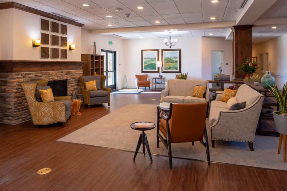 A cozy senior living facility common area with a stone fireplace on the left, two patterned armchairs with yellow cushions, a beige sofa, a gray sofa, and a brown leather chair arranged around a beige rug. The room has wooden flooring, a wooden ceiling beam, wall sconces, framed artwork on the walls, and various plants and decorative items. There is a glass door leading outside in the background.