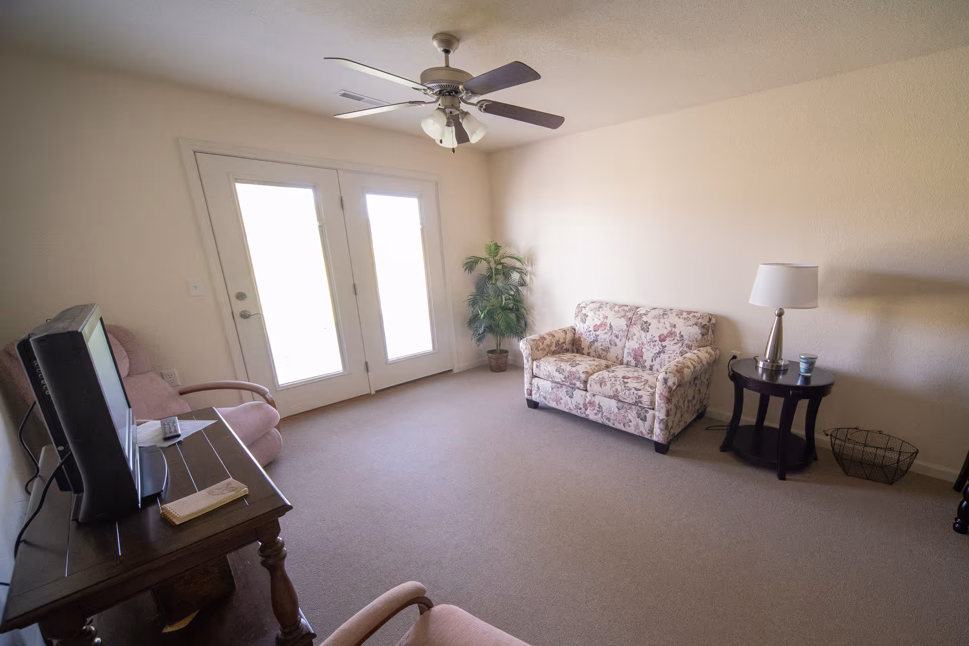 A small living room with beige walls and carpet, featuring a floral-patterned loveseat, a round side table with a lamp and a cup, a potted plant in the corner, two armchairs, a wooden table with a television and remote controls, and double glass doors letting in natural light.