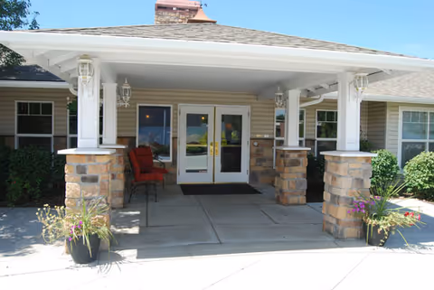 Entrance to a building with a covered porch supported by stone and white pillars, two red chairs on the left side, potted plants on either side of the entrance, and double glass doors in the center.