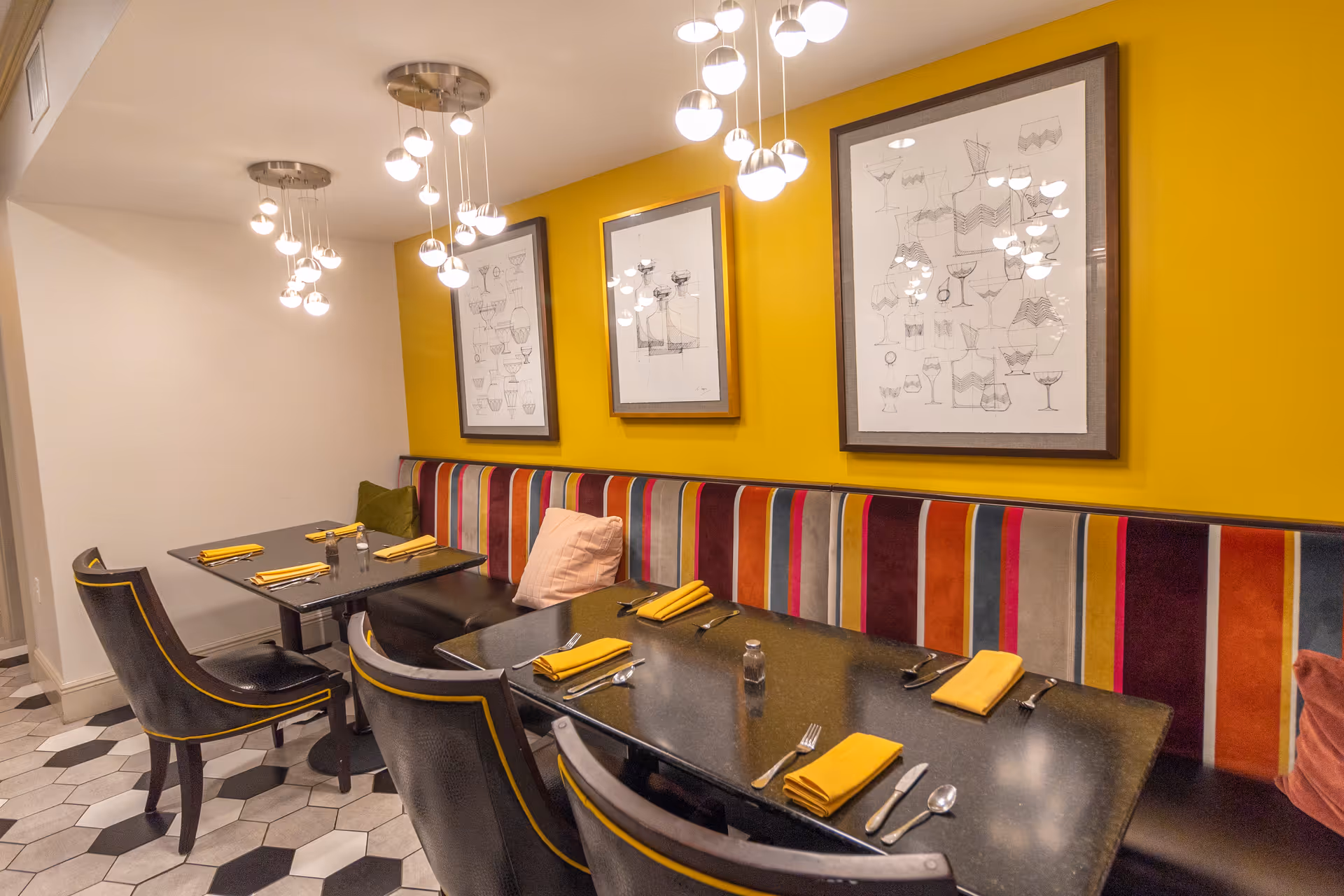 A dining area with two black tables set with yellow napkins, silverware, and salt and pepper shakers. The seating includes black chairs with yellow trim and a long bench with a colorful striped backrest and pillows. The wall behind the bench is painted yellow and decorated with three framed abstract art pieces. Modern pendant lights with multiple round bulbs hang from the ceiling. The floor has a hexagonal tile pattern in black, white, and gray.