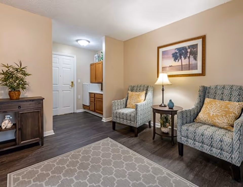 Small living room with two patterned armchairs flanking a round side table and lamp, a console table and rug, and an entryway/kitchen visible in the background.