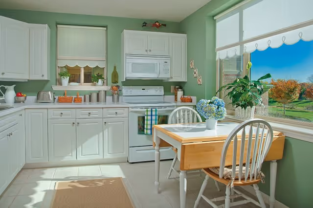A bright kitchen with white cabinets, a white stove and microwave, and a small wooden table with two chairs. The walls are painted green, and there is a large window showing a sunny outdoor scene with trees and grass. The table has a vase with blue flowers and a potted plant on the windowsill.