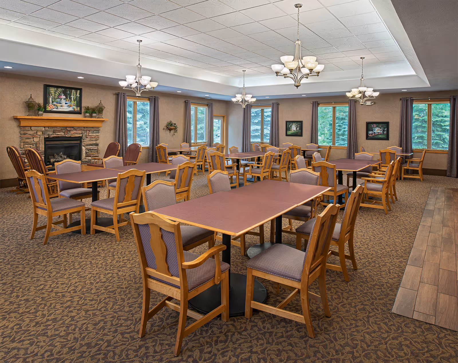 Spacious dining room with multiple rectangular tables and wooden chairs, chandeliers, a stone fireplace, and windows showing trees.