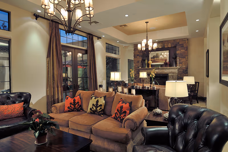A cozy living room with a brown sofa adorned with decorative pillows, two black leather armchairs, a wooden coffee table with a potted plant, and a stone fireplace in the background. The room is warmly lit by table lamps and a chandelier, with large windows and curtains letting in natural light.