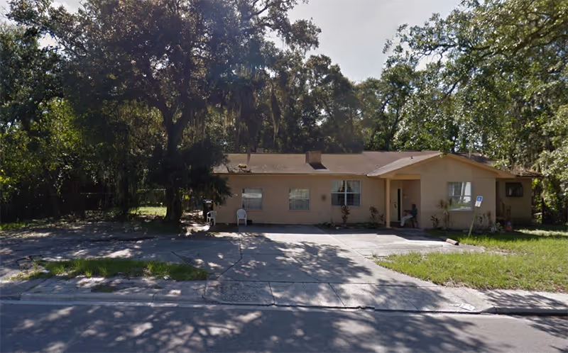 Single-story beige residential building with a sloped roof, surrounded by trees and greenery. There are two white chairs on the concrete driveway in front of the building, and a person sitting near the entrance. The area is shaded by large trees, and the building appears to be in a quiet neighborhood.
