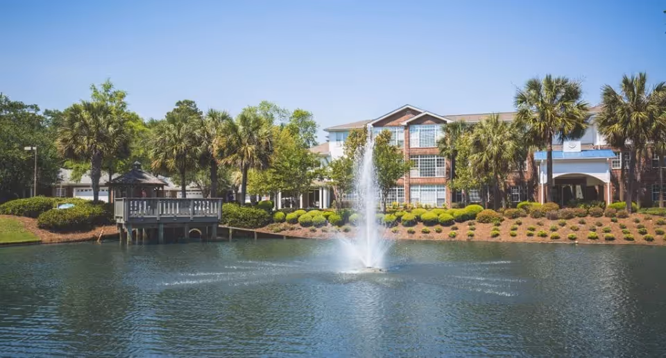 A scenic view of a senior living facility named The Lakes at Litchfield featuring a large pond with a water fountain in the center, surrounded by palm trees and well-maintained landscaping. In the background, there is a multi-story brick building with large windows and a covered entrance.