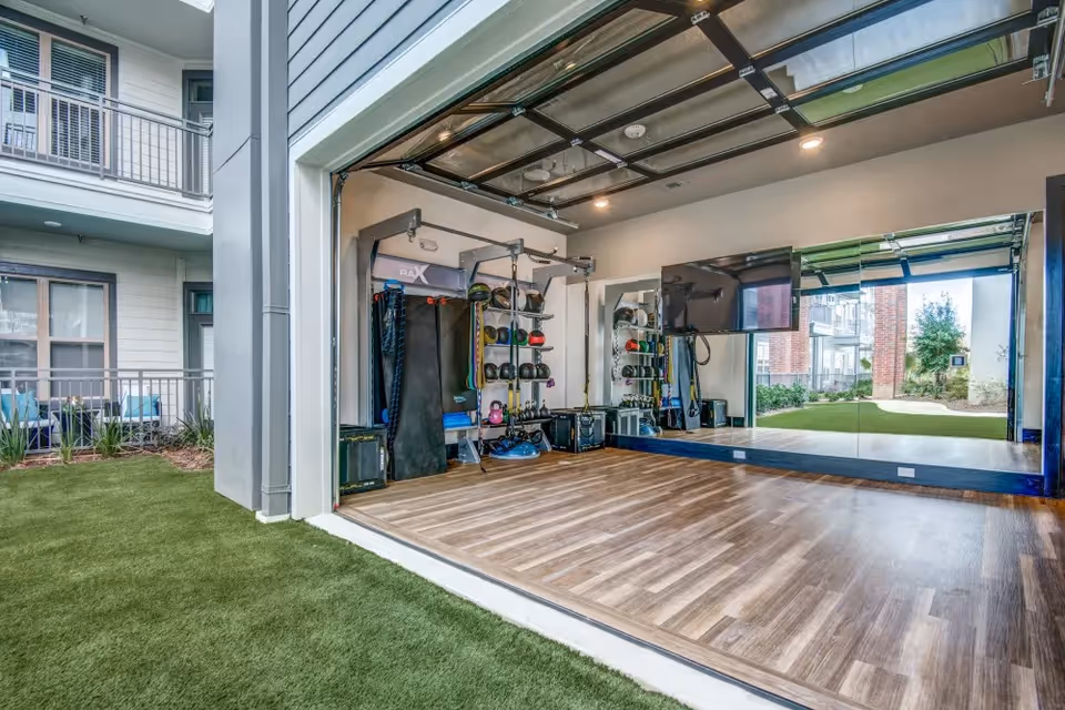 Open garage-style fitness studio with exercise equipment, a mirrored wall and TV, wood-look flooring, and an opening onto artificial turf.