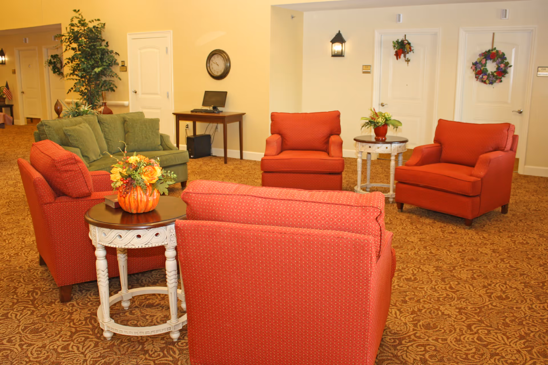 A cozy seating area in a senior living facility with four red armchairs arranged around two small round tables, one with a floral arrangement in a pumpkin-shaped vase. There is also a green sofa in the background, a small wooden desk with a computer, a wall clock, and two doors decorated with wreaths. The room has warm yellow walls and patterned carpet flooring.