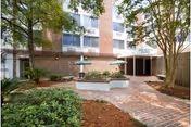 Brick-paved courtyard with raised planters, umbrella-shaded benches and trees in front of a multi-story residential building entrance.
