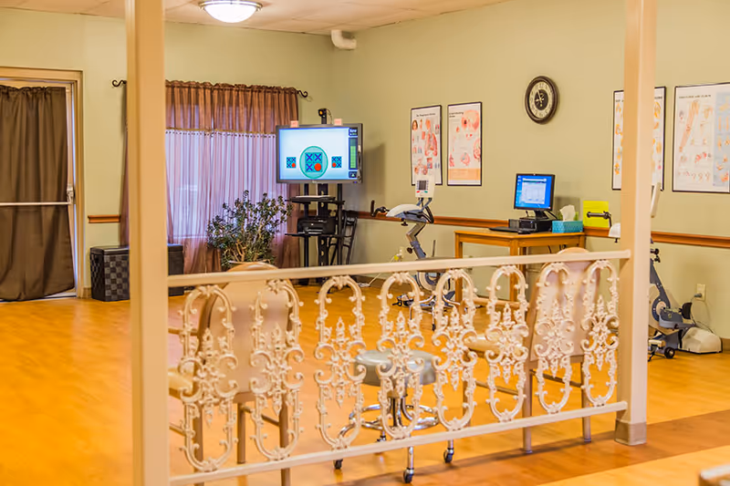 Interior view of a rehabilitation room with wooden flooring, exercise equipment including stationary bikes, a TV screen displaying a tic-tac-toe game, anatomical charts on the walls, a clock, and decorative white railing in the foreground.
