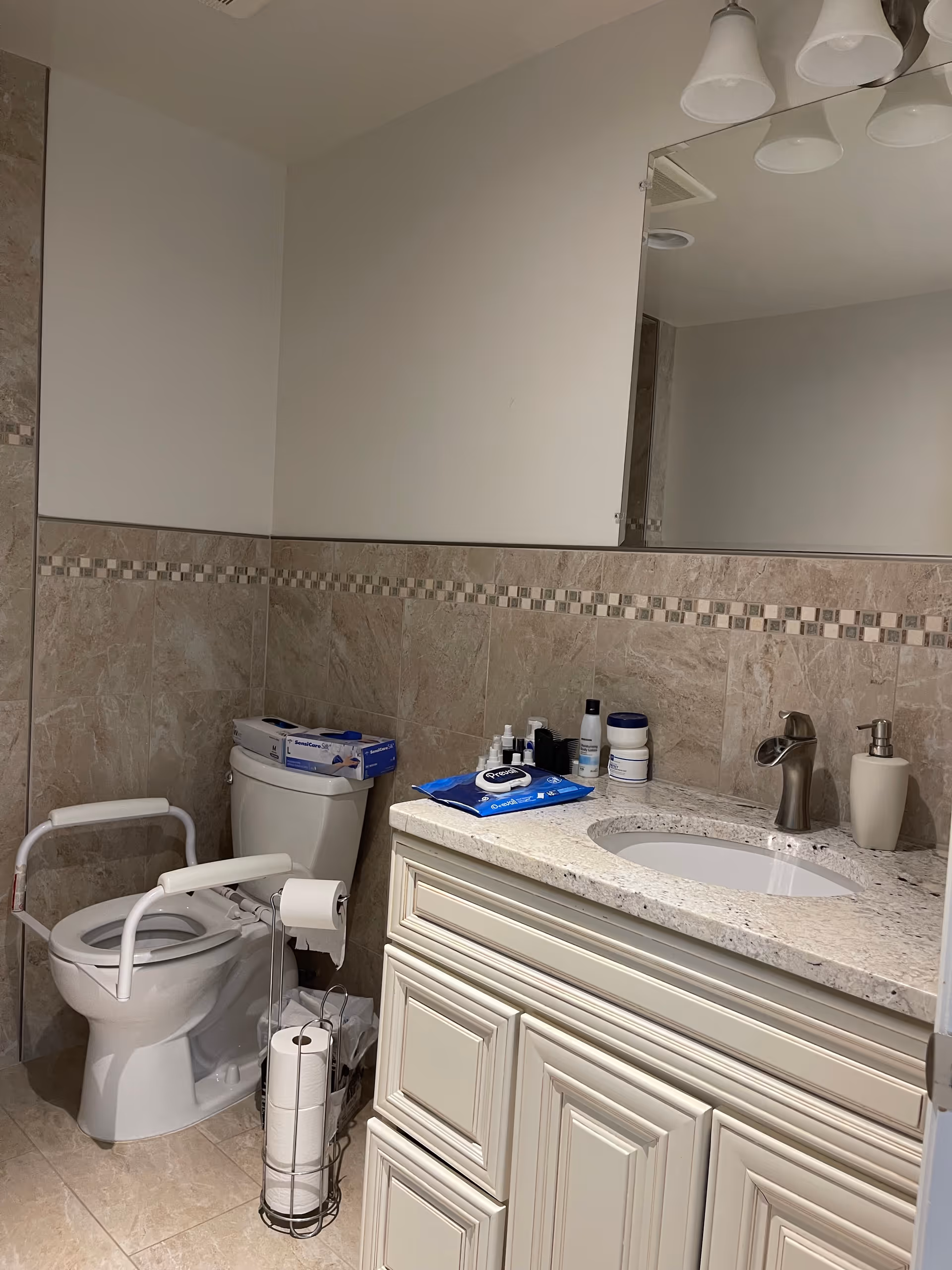 A bathroom with beige tiled walls and floor, featuring a toilet with white safety rails, a toilet paper holder with extra rolls, and a countertop with a sink, soap dispenser, and various personal care items. A large mirror and overhead light fixtures are visible above the sink.