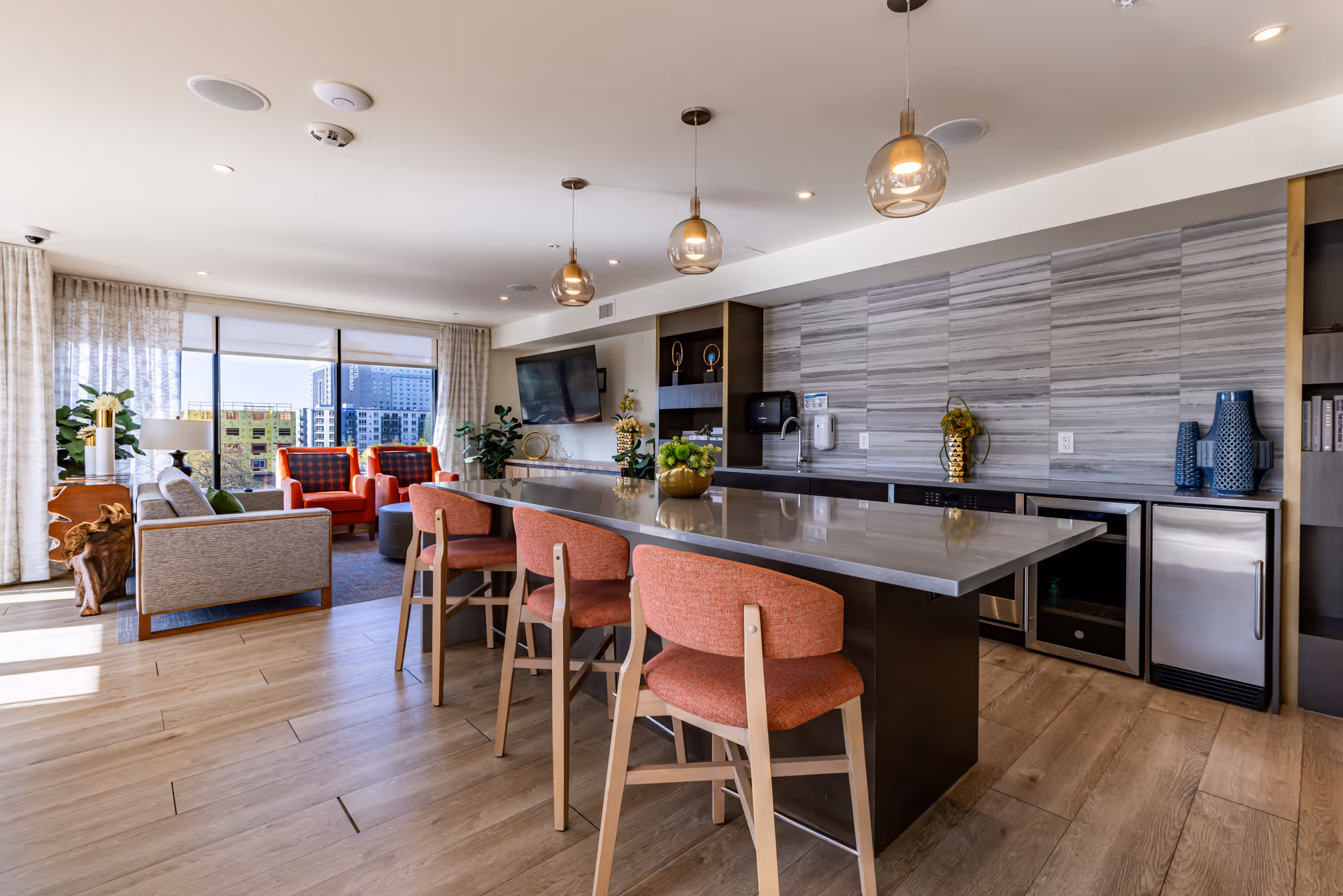 Open-plan communal living area and kitchenette with a large island, coral bar stools, seating area with sofas and red armchairs, and city views through floor-to-ceiling windows.