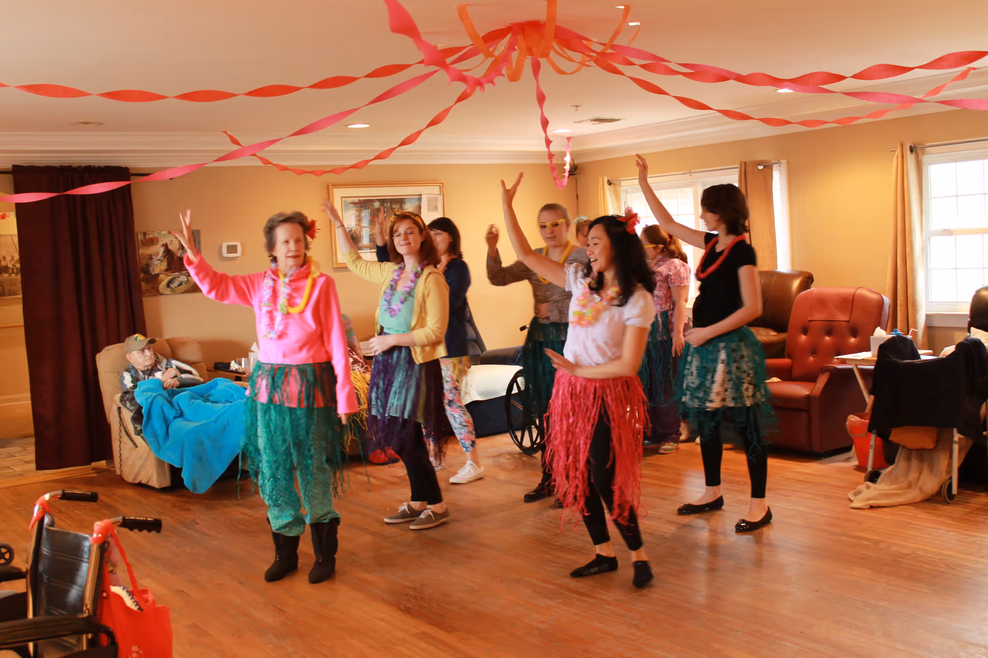 A group of women dressed in colorful Hawaiian-themed outfits with grass skirts and leis are dancing in a living room decorated with red and pink streamers. In the background, an elderly person is seated in a recliner covered with a blue blanket. The room has wooden flooring, comfortable chairs, and large windows with curtains.