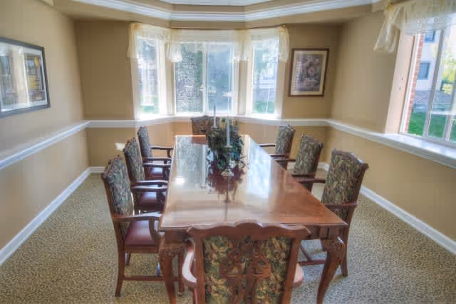 A dining room with a long wooden table surrounded by eight upholstered chairs with floral patterns. The room has beige walls with white trim, carpeted floor, and three large windows with sheer white curtains letting in natural light. There are framed pictures on the walls and a decorative centerpiece on the table.