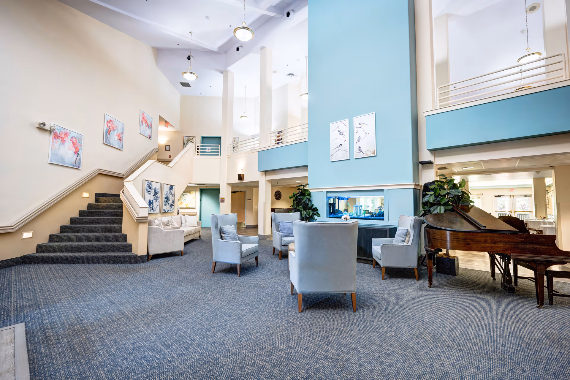 A spacious and well-lit common area in a senior living facility featuring a seating arrangement with several light gray armchairs around a built-in aquarium. The room has high ceilings, light beige walls, and a blue accent wall with framed artwork. There is a grand piano on the right side and a staircase with carpeted steps on the left. The area has carpeted flooring and modern hanging light fixtures.