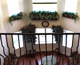 View from an upper level looking down into a bright sitting area with large windows, potted plants, and a round glass table surrounded by chairs. There are green plants on window ledges and a clock on the wall between the windows.