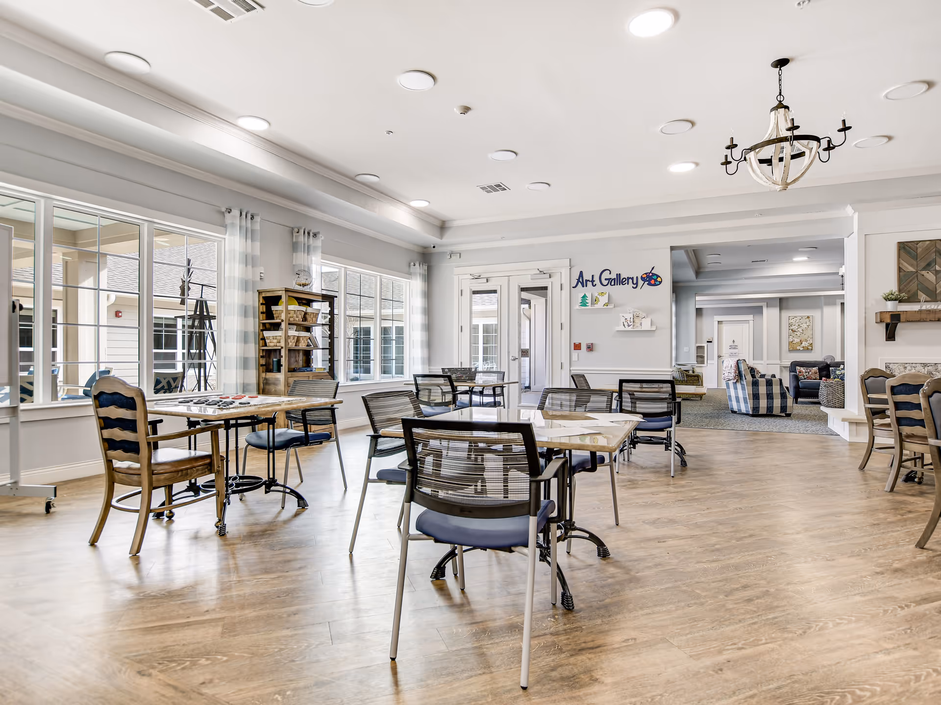 Bright and spacious interior room with multiple tables and chairs arranged for activities or dining. Large windows with sheer curtains allow natural light to fill the space. A sign on the wall reads 'Art Gallery' with colorful decorations. In the background, there is a cozy seating area with armchairs and a fireplace.