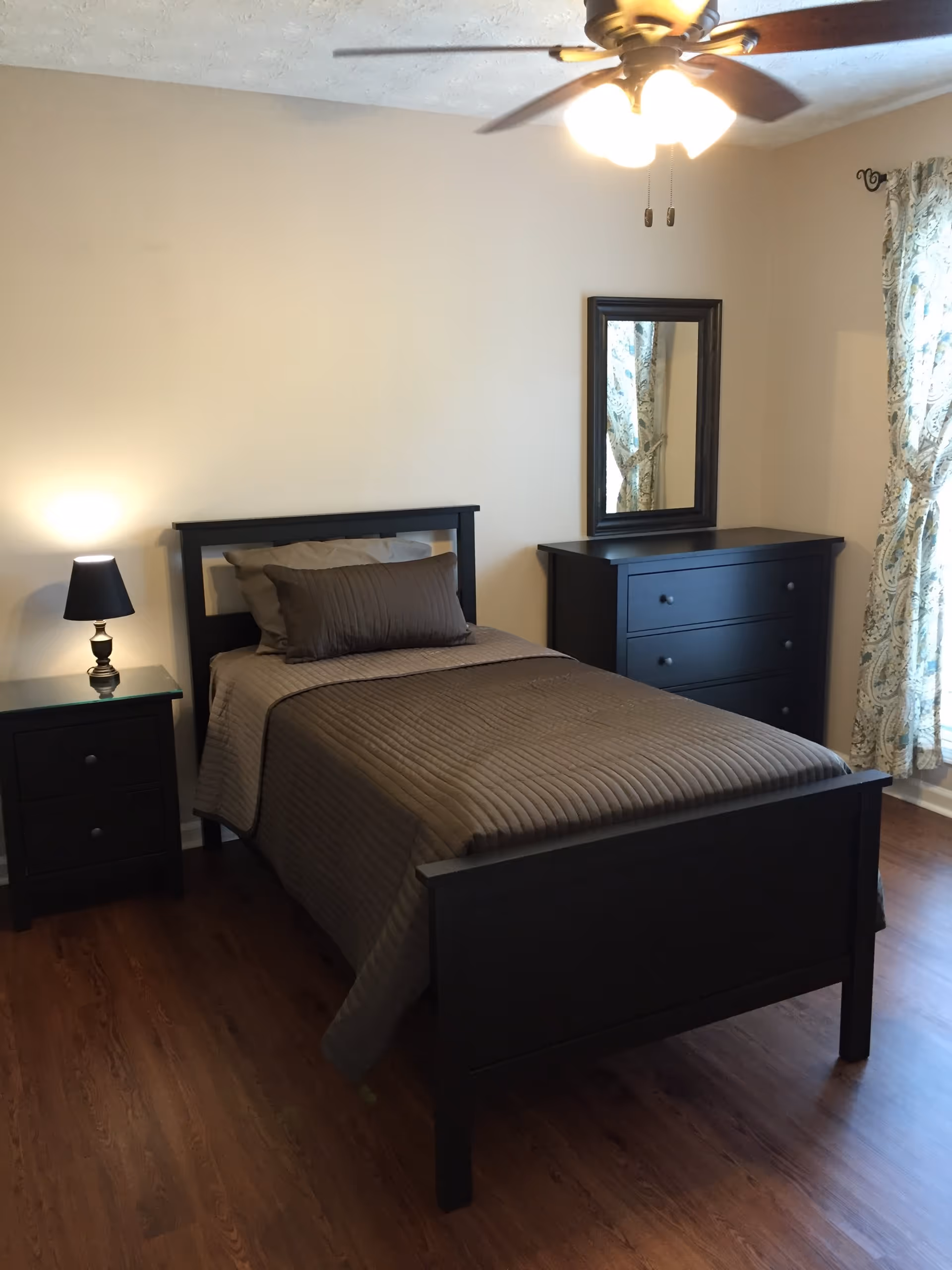 A bedroom with a single bed featuring a dark wooden frame and brown bedding. Next to the bed is a matching dark wooden nightstand with a small lamp on it. Across from the bed is a dark wooden dresser with a mirror above it. The room has wooden flooring, beige walls, a ceiling fan with lights, and a window with patterned curtains.