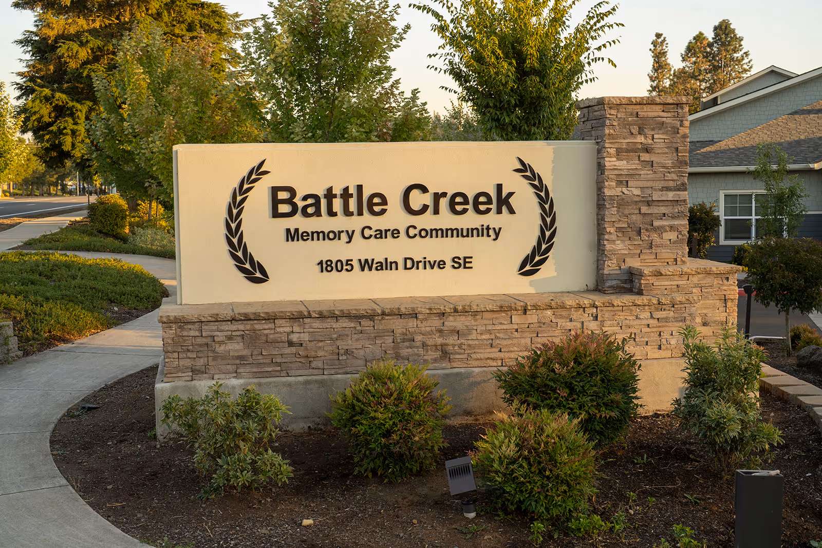 Outdoor view of a stone and stucco sign for Battle Creek Memory Care Community located at 1805 Waln Drive SE, surrounded by landscaping with bushes and trees along a sidewalk.