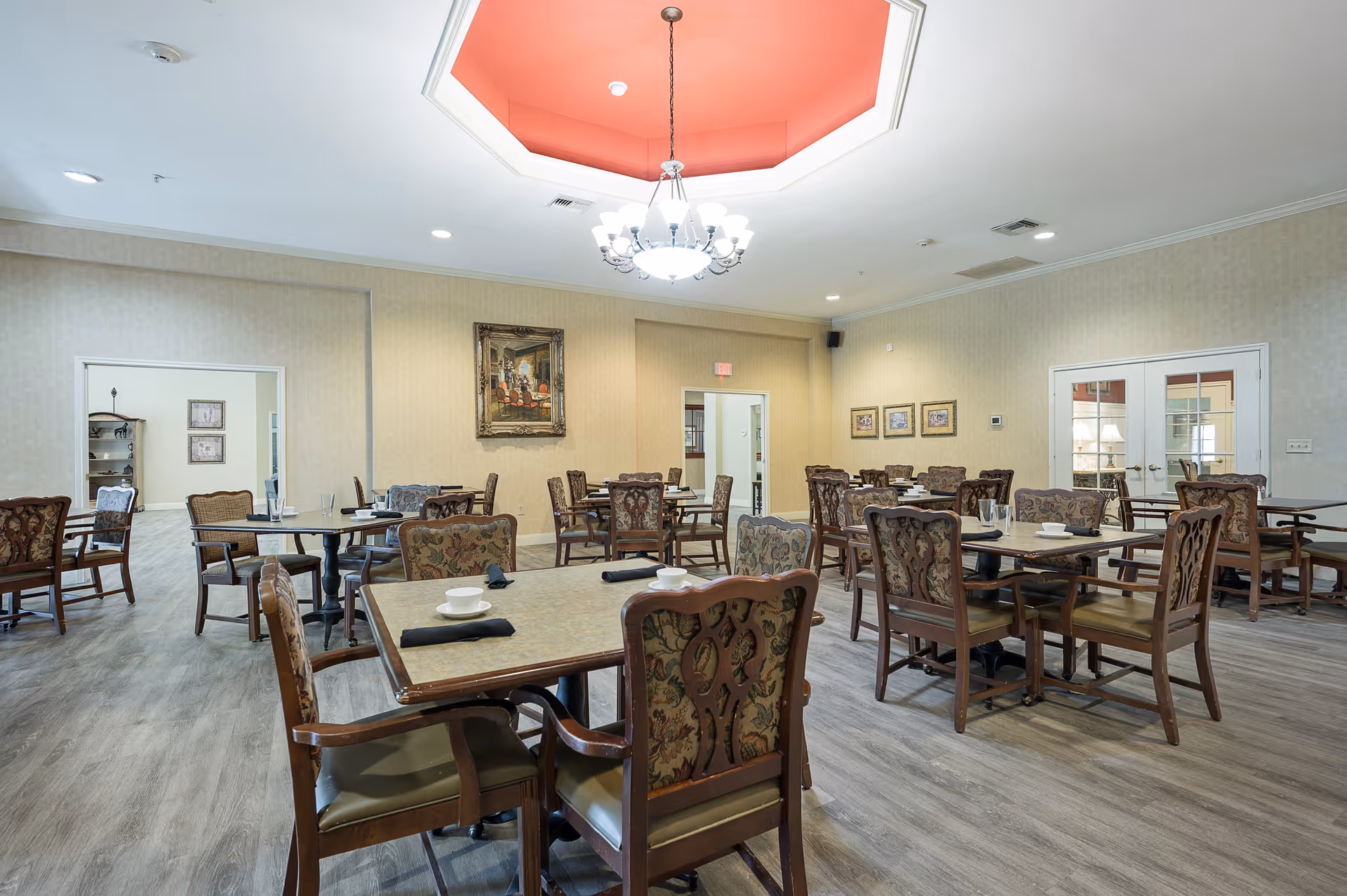 A spacious dining room with multiple wooden tables and chairs arranged neatly. Each table has a cup and a black napkin. The room features a decorative chandelier hanging from a recessed ceiling with a red accent. The walls are adorned with framed paintings, and there are doorways leading to other rooms.
