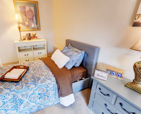 A cozy bedroom featuring a bed with blue and brown bedding, multiple pillows, a tray with folded towels on the bed, a white dresser with a lamp and books, a framed picture on the wall, and a gray nightstand with a lamp and decorative items.