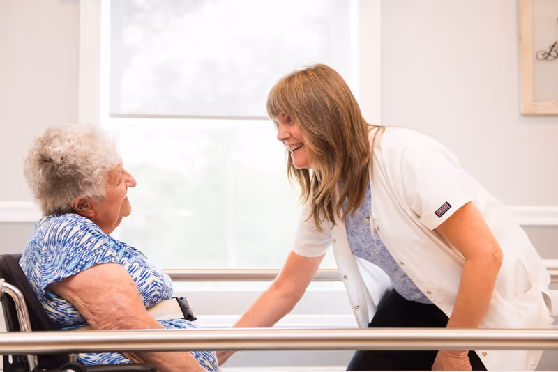 A healthcare worker in a white coat smiling and interacting with an elderly woman seated in a wheelchair in a bright room with a large window in the background.