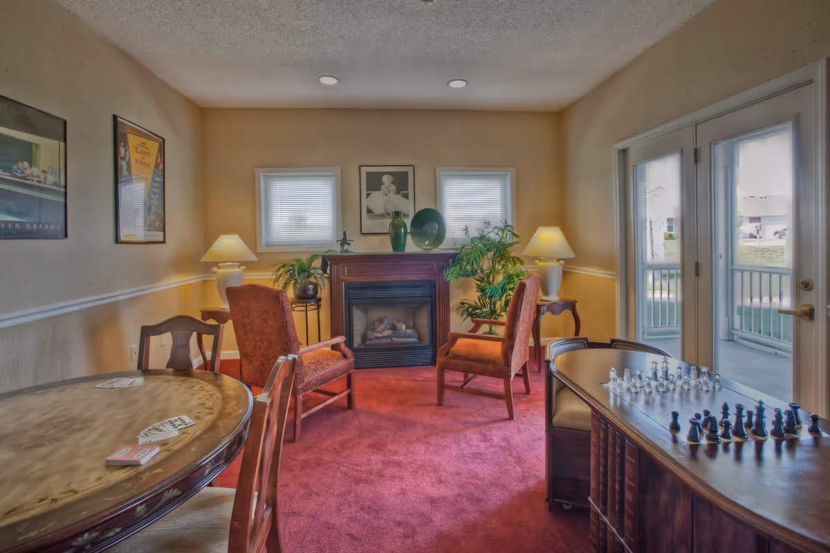 A cozy living room area with two upholstered armchairs facing a fireplace. The room has a red carpet, two small windows above the fireplace, and two table lamps on side tables. There are framed pictures on the walls and a round wooden table with playing cards on it. A chessboard with chess pieces is set up on a wooden table near glass doors leading outside.