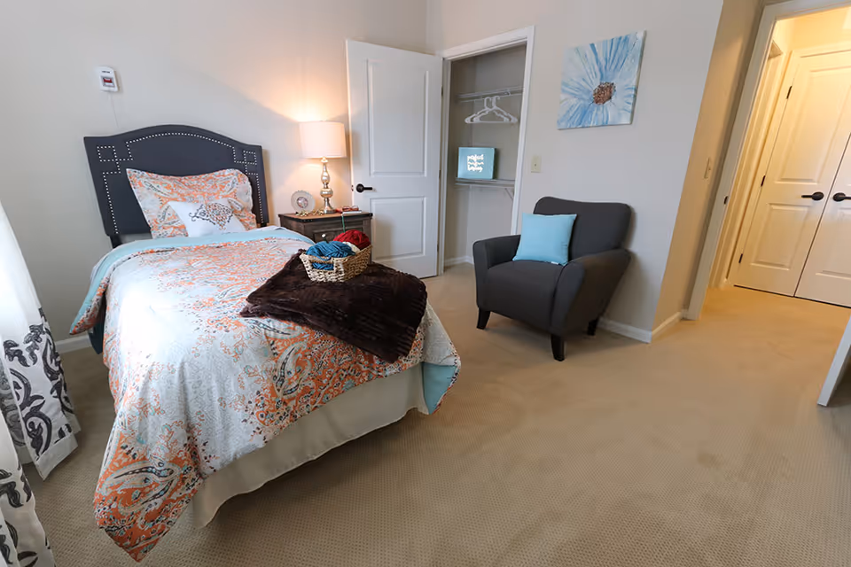 A bright bedroom with a single bed featuring patterned bedding, a bedside lamp, a gray armchair, and an open closet.