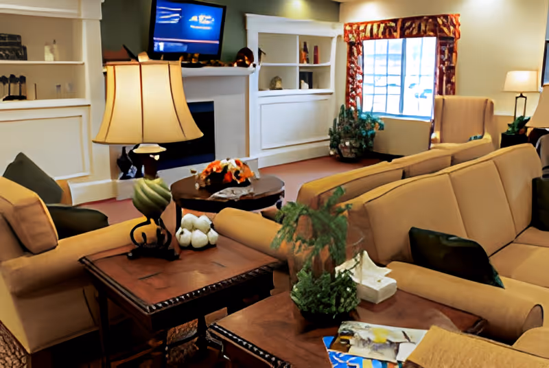 A senior living common room with beige sofas and armchairs, wooden side tables and lamps, a TV over a fireplace, and plants by a window.