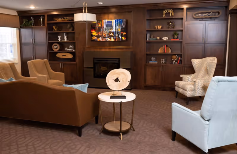 A comfortable communal living room with upholstered chairs and sofa arranged around a small round table, in front of built-in wooden shelving and a fireplace.