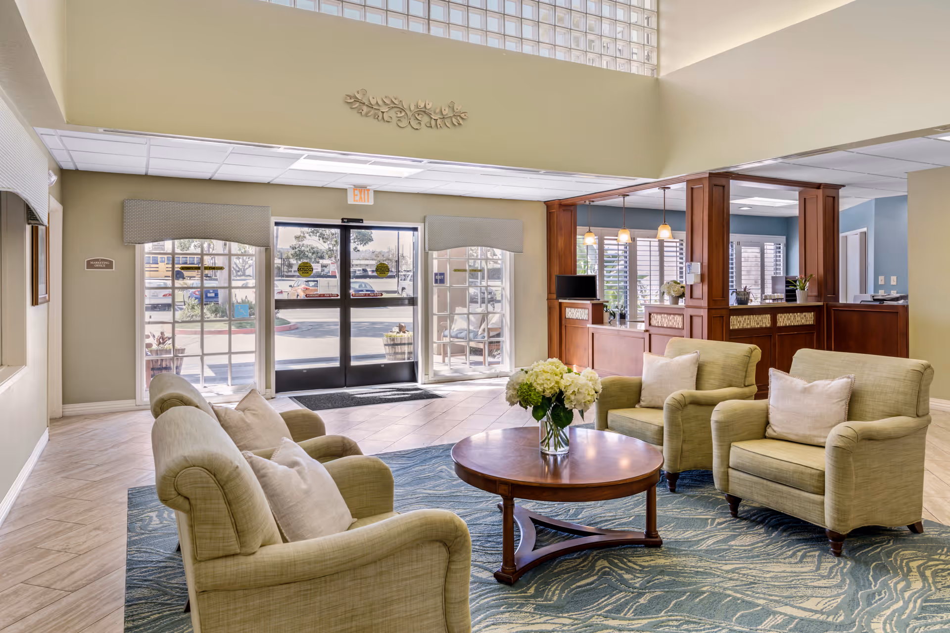 A bright and welcoming senior living facility lobby with beige armchairs and a round wooden coffee table with a vase of white flowers. The area has large windows and glass doors letting in natural light, a blue patterned rug, and a wooden reception desk with pendant lights in the background.