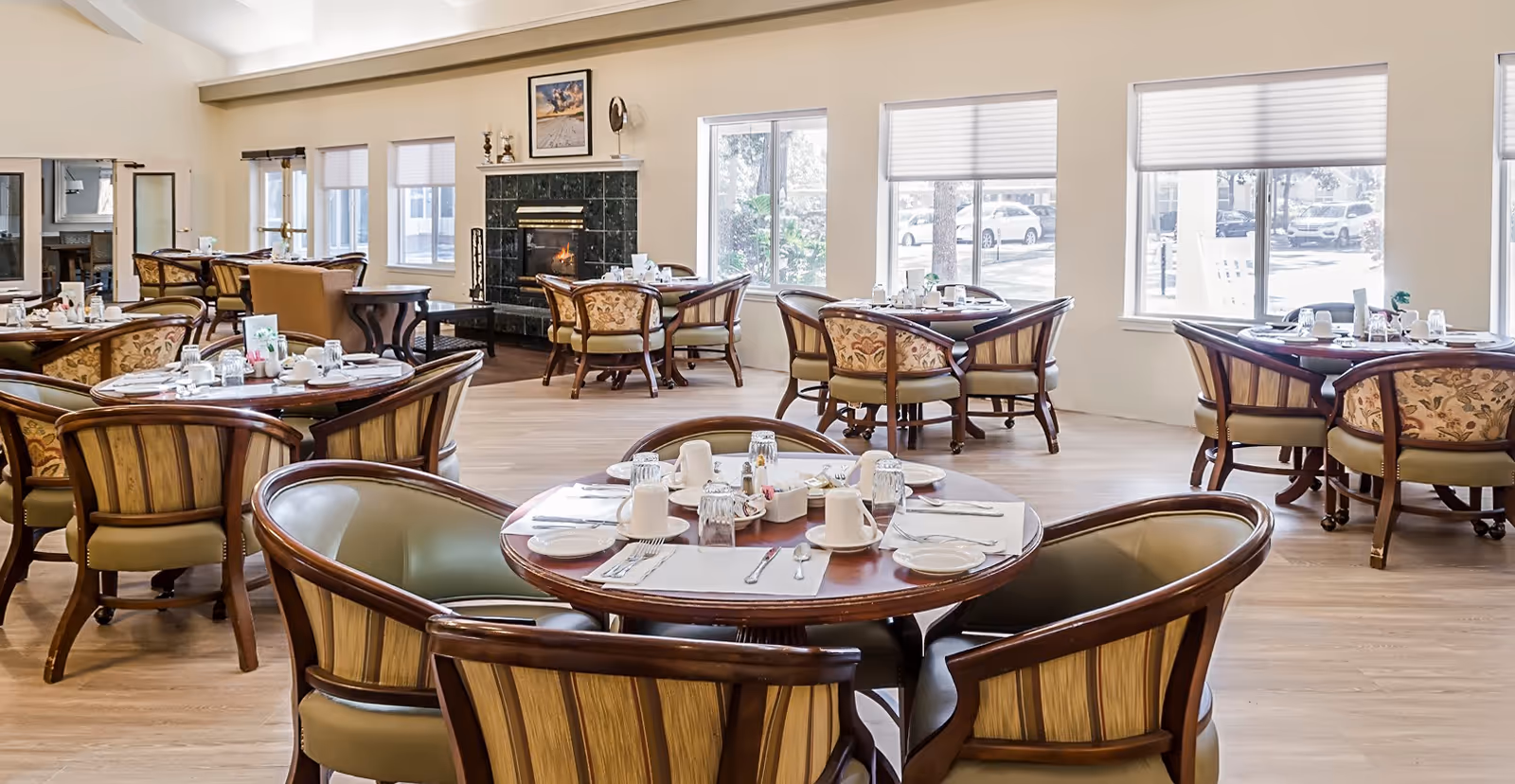 A bright dining room with multiple round wooden tables set with white cups, plates, and silverware. The room features large windows with blinds, a fireplace with a black marble surround, and comfortable upholstered chairs around each table. Light wood flooring and neutral-colored walls create a warm and inviting atmosphere.
