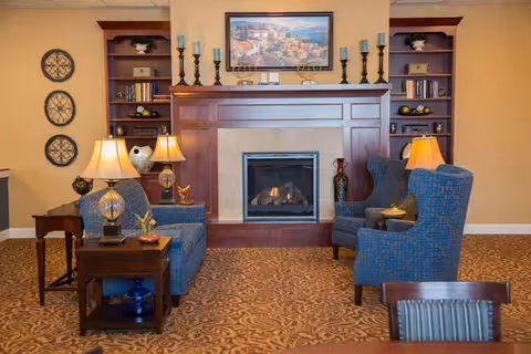 A cozy senior living common area featuring a lit fireplace with a wooden mantel, flanked by built-in bookshelves. Two blue upholstered armchairs and a matching loveseat are arranged around the fireplace, accompanied by side tables with lamps. The walls are decorated with framed artwork and decorative plates, and the carpet has an intricate pattern.