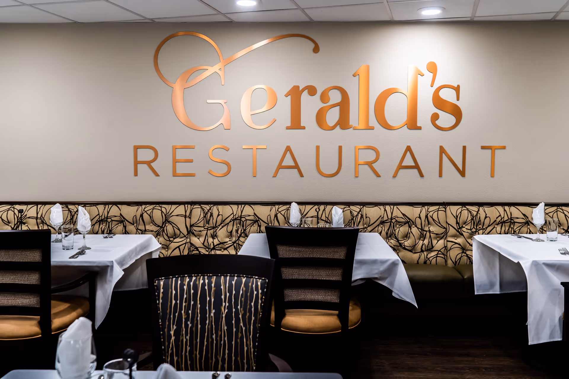 Interior view of Gerald's Restaurant with tables set with white tablecloths, napkins, glasses, and cutlery. There is a patterned cushioned bench along the wall and wooden chairs with cushioned seats. The restaurant name 'Gerald's RESTAURANT' is displayed prominently on the wall in large orange letters.