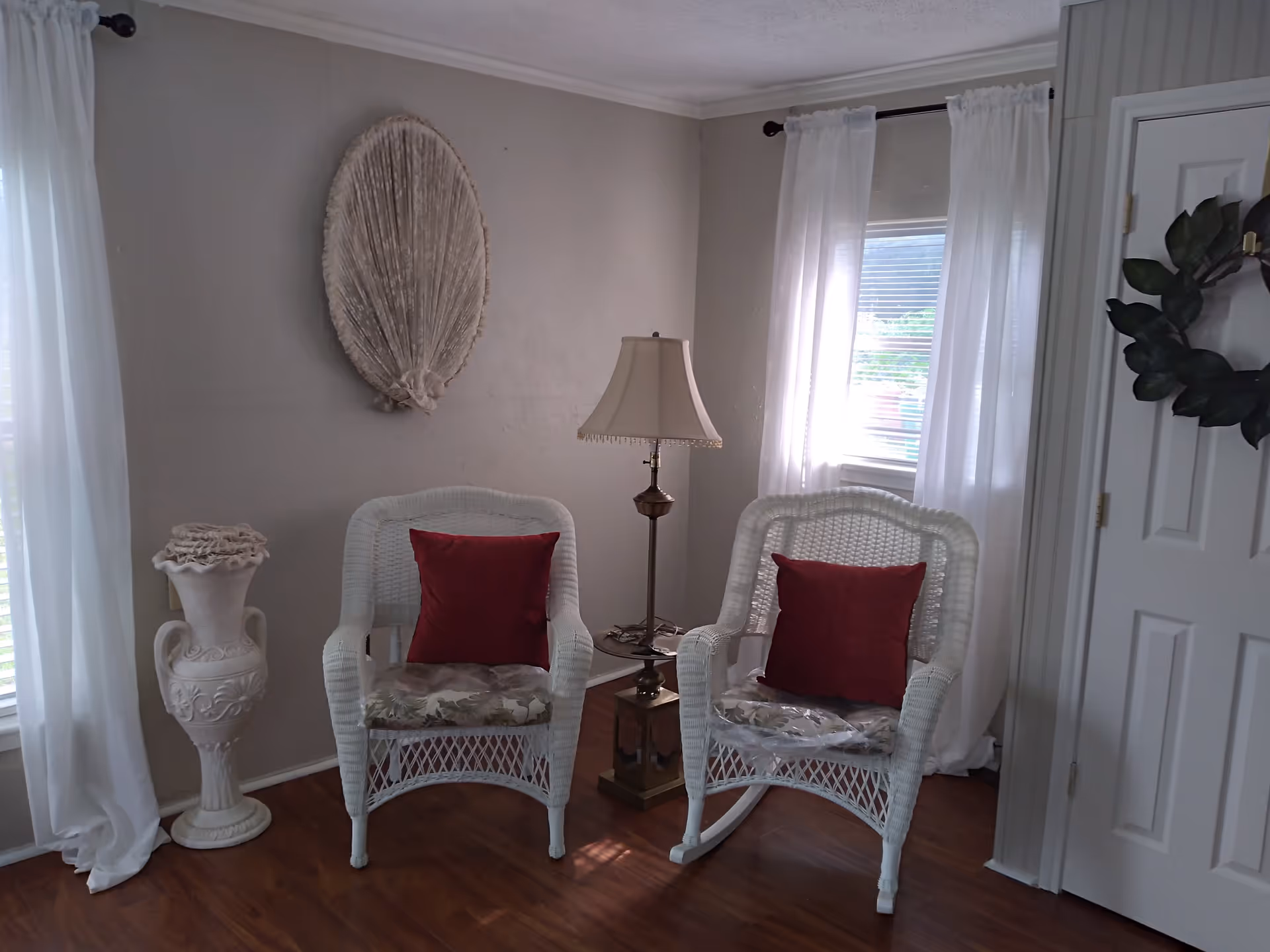 A cozy corner of a living room with two white wicker chairs, each with a red cushion and floral seat cushion. Between the chairs is a brass floor lamp with a beige lampshade. The walls are light-colored, with a decorative large fan-shaped wall hanging above one chair and a white vase with a ruffled top on the floor. White sheer curtains cover two windows, and a white door with a green wreath is visible on the right side.