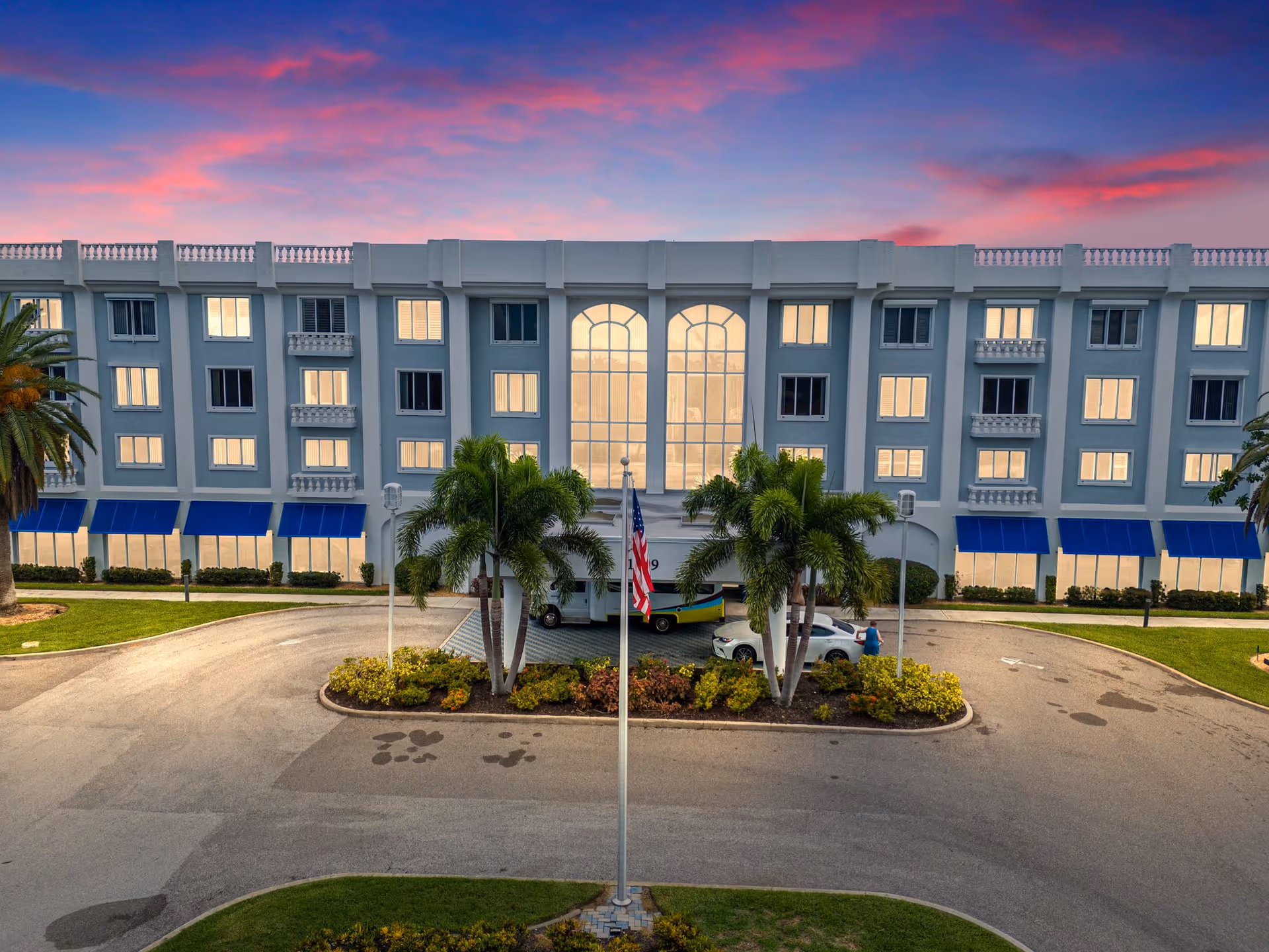 Front exterior view of The Summit of Fort Myers building at dusk with a vibrant pink and purple sky. The building has multiple windows with blue awnings on the ground floor, palm trees, an American flag on a flagpole, and a driveway with parked cars.