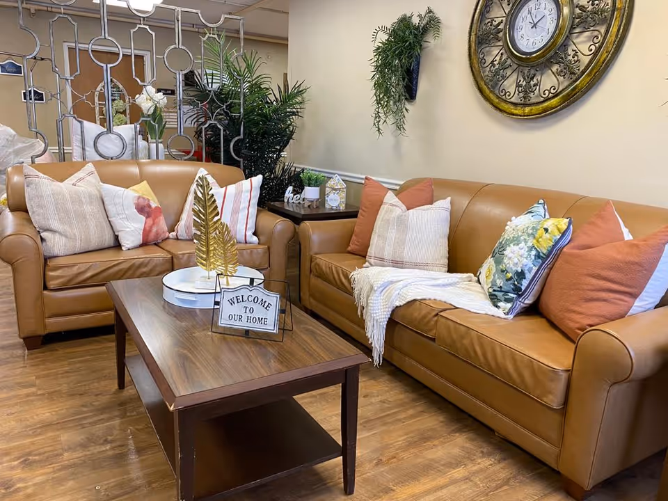 A cozy living room area with two tan leather sofas adorned with various decorative pillows and a white throw blanket. A wooden coffee table sits in front of the sofas with a decorative golden leaf sculpture and a small sign that reads 'WELCOME TO OUR HOME'. The wall behind the sofas features a large ornate clock and a hanging plant. There is a decorative metal room divider and some greenery in the background.