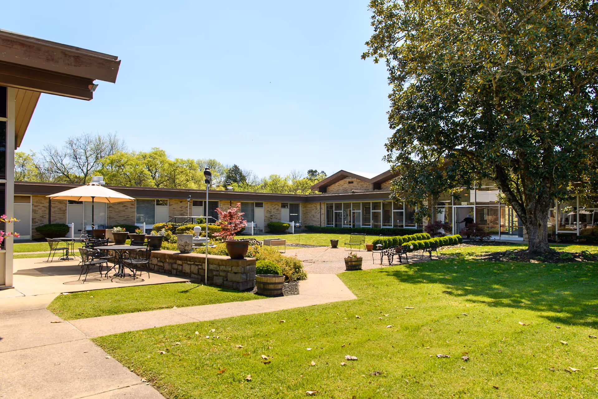 Outdoor courtyard area of Creekside Center for Rehabilitation & Healing featuring green grass, a large tree, patio tables with umbrellas, benches, potted plants, and a single-story building with large windows in the background under a clear blue sky.